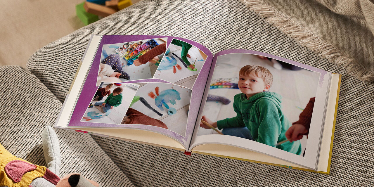 Auf einem Sofa liegt ein geöffnetes CEWE FOTOBUCH neben einem Kuscheltier und einer Decke. Auf der linken Seite sind vier Fotos zu erkennen, die sich leicht überlappen. Alle zeigen ein Kind, das mit den Farben aus einem Tuschkasten ein Bild malt. Die rechte Seite zeigt ein grosses Foto von einem Jungen, der in die Kamera schaut. Die Doppelseite ist mit einem violetten Aquarellmuster hinterlegt.