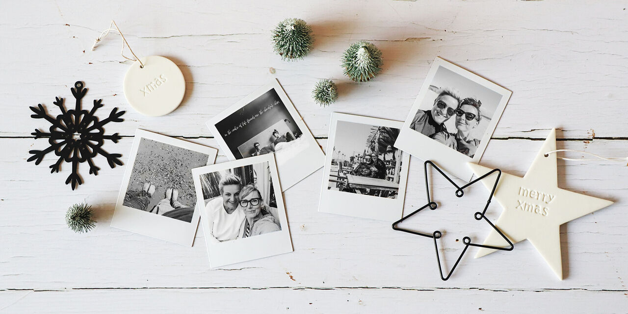 Sur une table, il y a plusieurs magnets photo avec des motifs photo en noir et blanc de deux amies en vacances. Tout autour, il y a quelques pendentifs de Noël, également en noir et blanc.