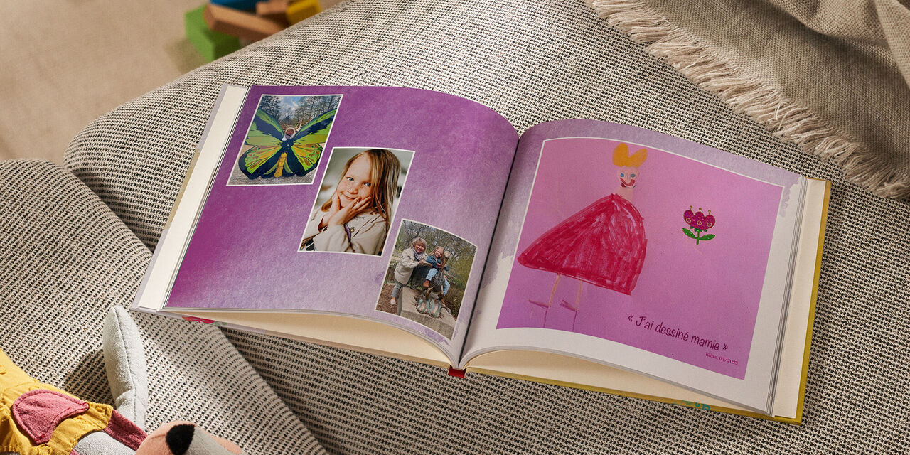 Sur un sofa, un LIVRE PHOTO CEWE ouvert est posé à côté d’un animal en peluche et d’une couverture. Sur la page de gauche, trois photos sont disposées en diagonale. La première photo montre de grandes ailes de papillon derrière lesquelles se tient une femme. Sur la photo centrale, on peut voir le portrait d’une fillette. Une troisième photo montre la fillette avec sa grand-mère sur une statue de pélican. La page de droite montre un dessin d’enfant représentant une femme et une fleur sur du papier rose. Le titre est le suivant  : « J’ai dessiné ma grand-mère. Elina, 03/2023 ». L’arrière-plan de la page du livre photo est décoré avec un motif d’aquarelle violet.